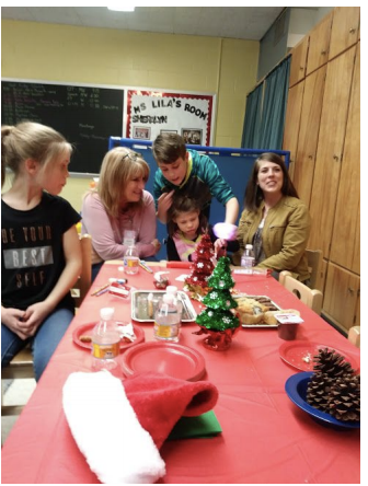 A student’s mother, brother, sister and grandmother are sitting/standing at a table decorated with holiday decor and trays of cookies/treats. The student’s brother is helping her to open her gift.