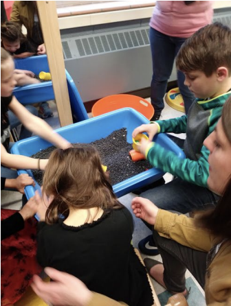 Children sitting on the floor with their hands exploring items in dirt in a plastic blue rectangular container.