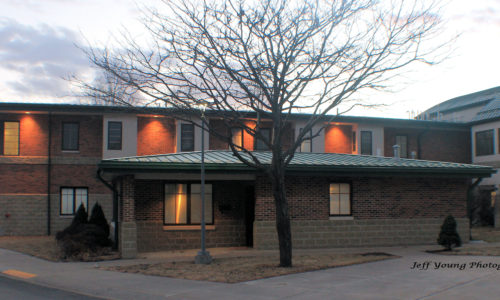 Two story brick building with a large tree, empty of leaves and a light pole in front. Night Lights beam from the 2nd story eaves shining orange in the sunset.