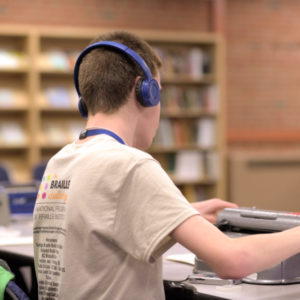 A Male Student With Back To Camera Sitting At A Table With A Braille Writer In Front. He Is Wearing Headphones And The 2019 Braille Challenge T-shirt. Bookshelves On The Wall In The Background.