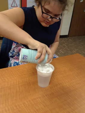 A student sitting at table with a clear plastic drinking cup. The student is squirting shaving cream into the liquid in the cup. Foam appears on top.