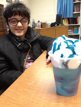 Student sitting at table with a plastic clear cup filled with blue liquid, topped with white foam and blue food coloring dripping on the foam.