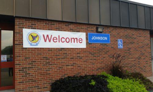 a close up of a red brick building with a printed white banner that says Welcome and the KSSB logo. A blue plastic large name plate that says Johnson.