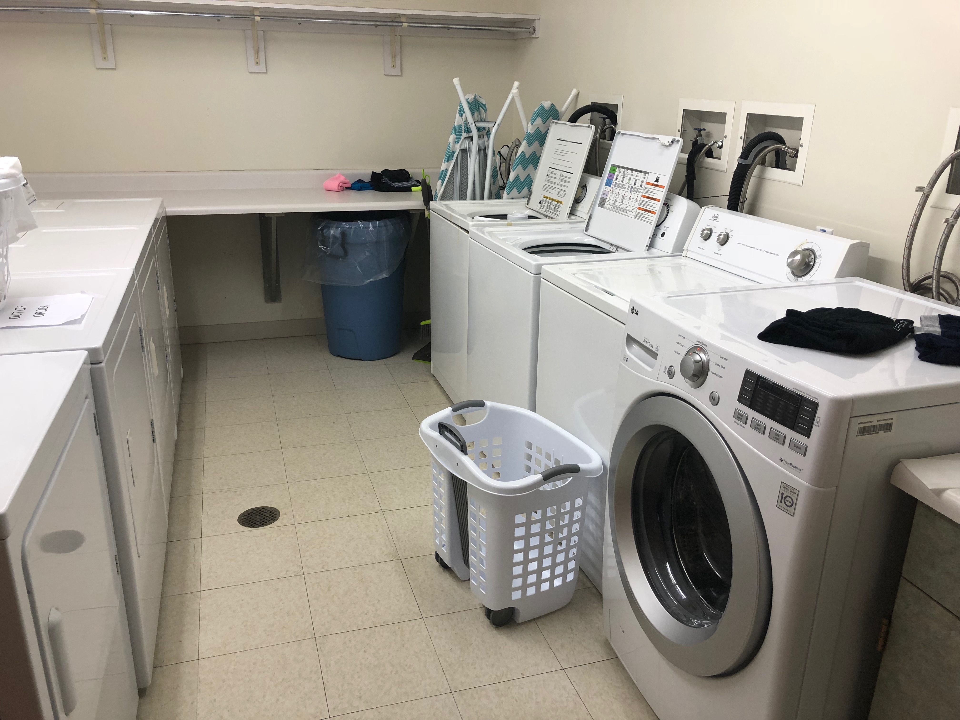 On the right is 4 washing machines. One is front loading and 3 are top loading. A laundry basket is on the floor in front of washer. Left side is 4 dryers. A long rectangular table runs along the back wall for folding clothes. 2 ironing boards are on right between washing machine and table.