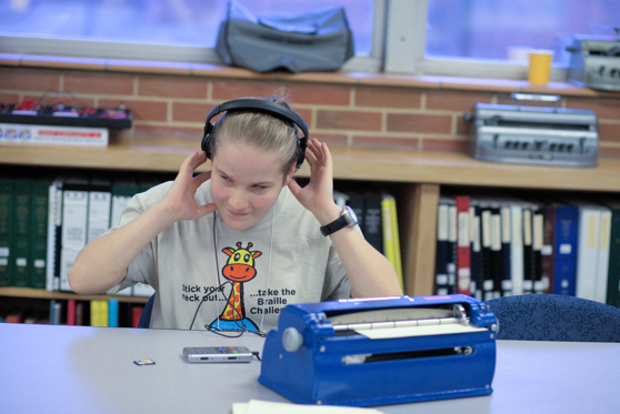 Student listening to headphones and a braille writer sitting on table in front of her.