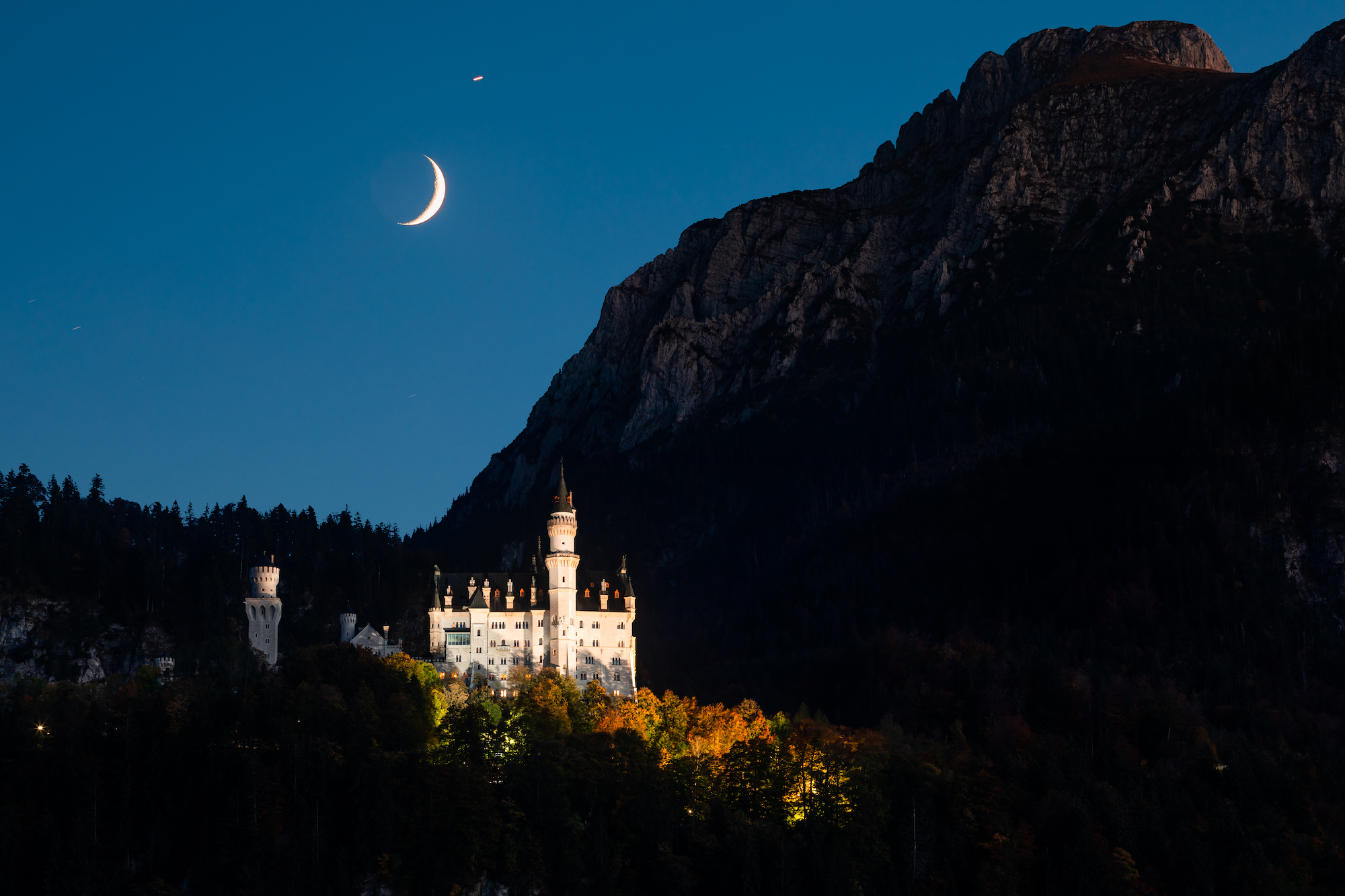 On the side of a mountain the moonlight shines on a large castle and surrounding trees with fall leaves.