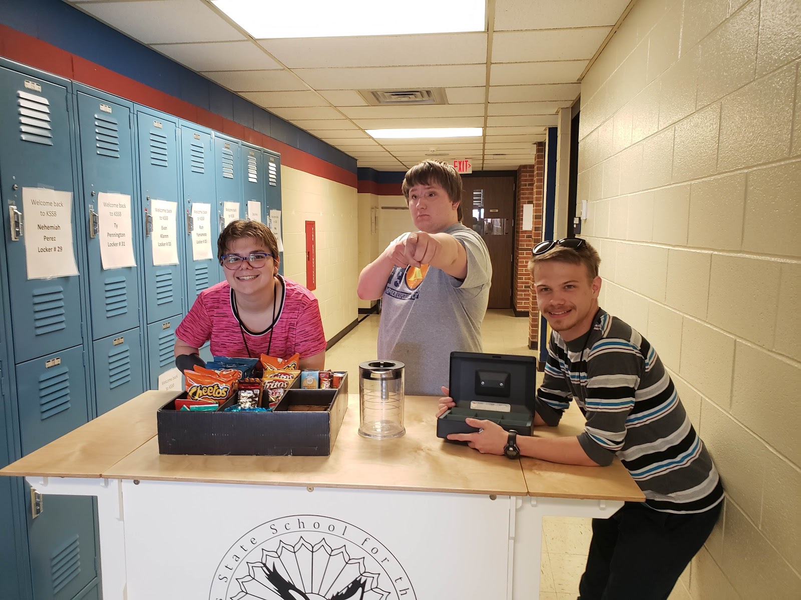 3 students standing at the Coffee Cart - one on the left, leaning over the snack basket. A second student behind the cart pointing at the camera. A third student with elbows on cart holding the cash box.