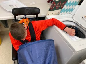 Student in a wheelchair holding a blue laundry basket on his lap and placing clothes into a washing machine.