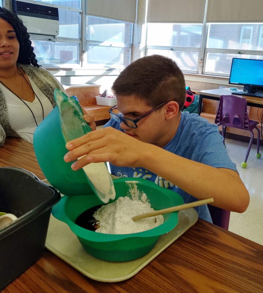 A student with hands on a round bowl, pouring flour into another bowl filled with moist ingredients.