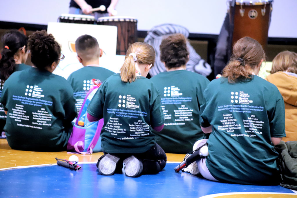 Severals students sitting on the floor, wearing the braille challenge t-shirt, with their backs facing the camera to show the sponsors listed on the back of the shirts. .