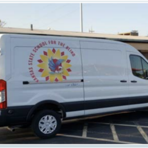 A Large White Van With KSSB’s New Logo Is Parked In The Circle Drive, Next To A Display Table Draped With A Cloth.