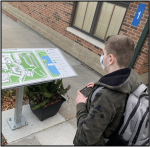 Student, holding a phone and standing in front of a Wayfinding Tactile Map located near entrance to building 1.