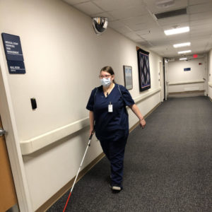 Student Wearing Navy Blue Scrubs, Backpack, Name Badge, And Holding A White Cane Walking Down An Empty Hallway