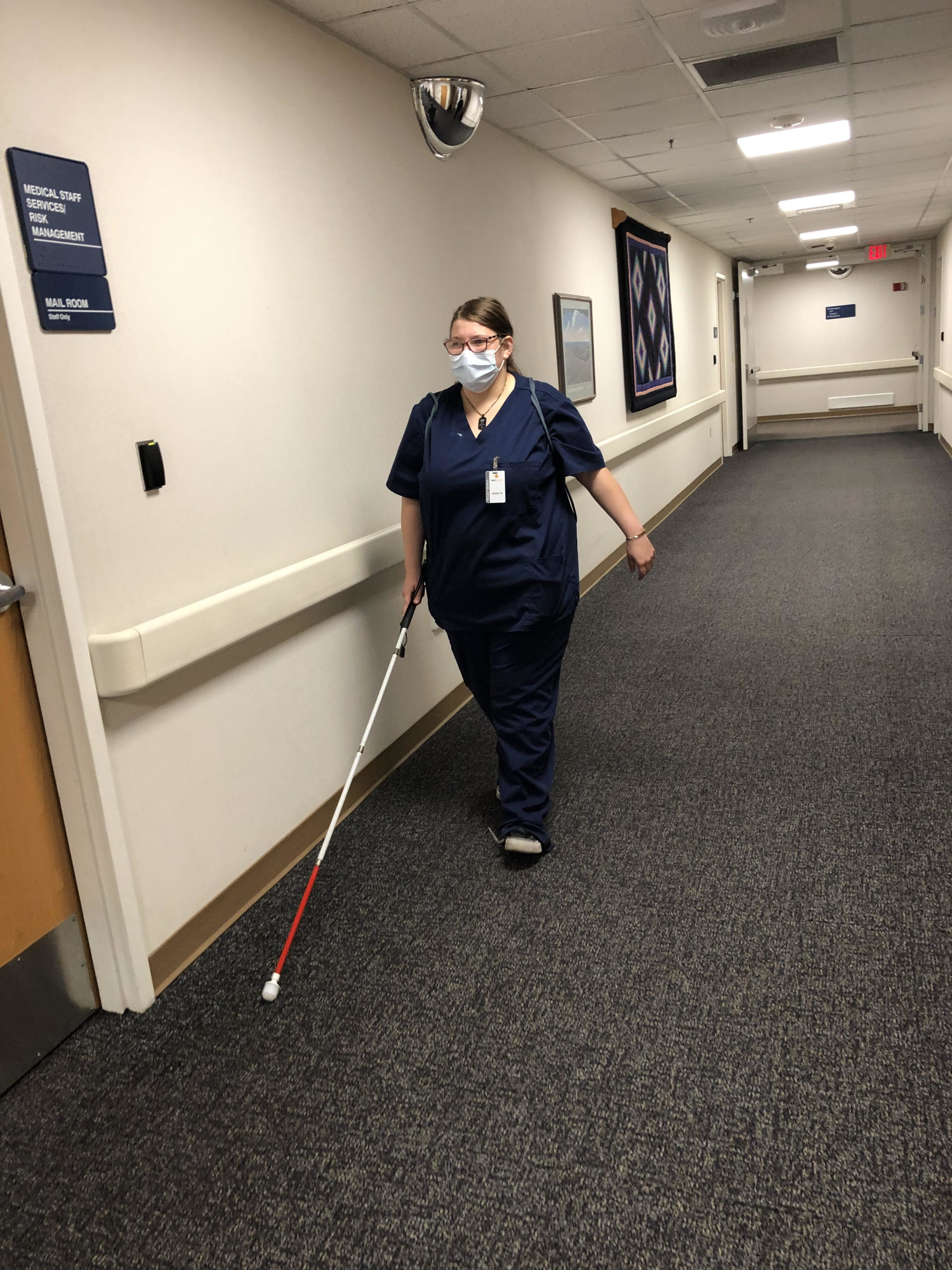 student wearing navy blue scrubs, backpack, name badge, and holding a white cane walking down an empty hallway