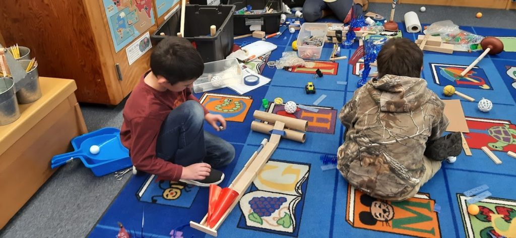 2 students sit on the floor among many materials creating a ramp for a marble
