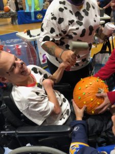 student in a wheelchair laughing while a teacher helps him hammer a golf tee into a pumpkin