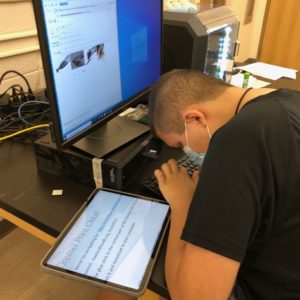 Student Sitting At A Computer Typing With Directions Enlarged On An IPad To His Left