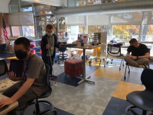 3 students working together in the makerspace. One is standing at a desk with a Braillenote, one is sitting at a desk holding an iPad, and the third is sitting in a chair with legs crossed listening