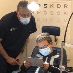 Boy Sitting In A Chair, Holding An Eye Chart With Light Overhead And Dr. Krug Looking On