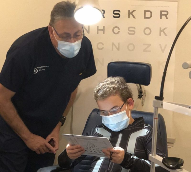 boy sitting in a chair, holding an eye chart with light overhead and Dr. Krug looking on