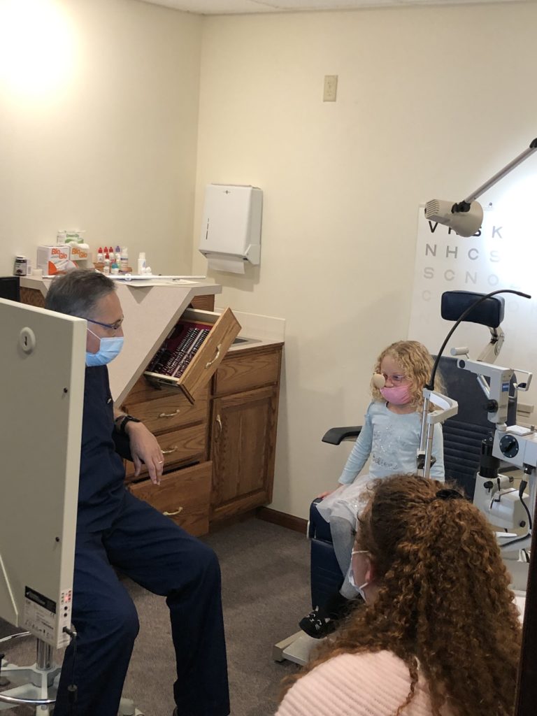 girl sitting in chair looking at a visual acuity chart with Dr. Krug and mom watching the girl