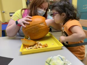 girl sticking her hand into a pumpkin while a teacher helps to hold the pumpkin