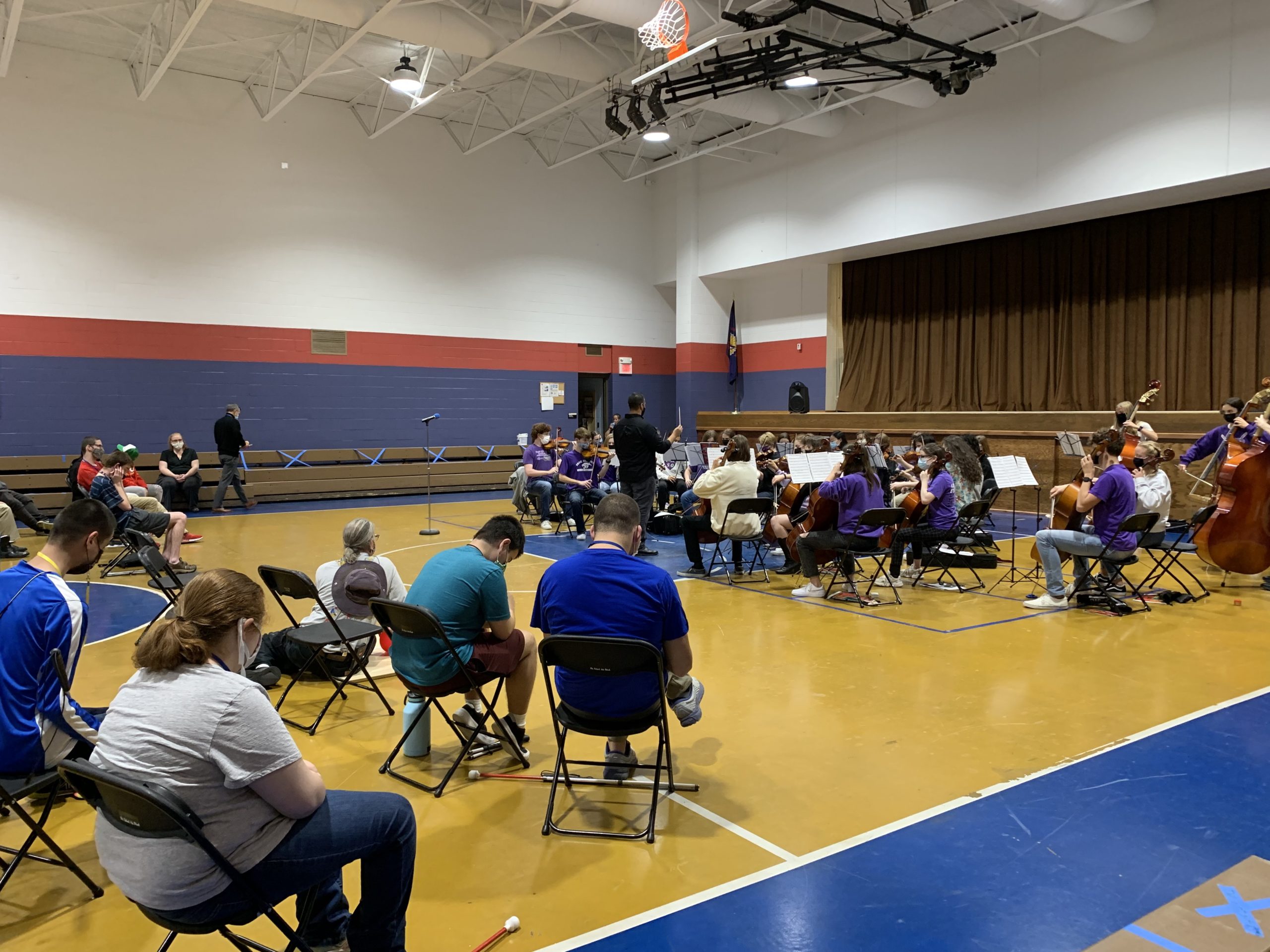 students and staff sit in chairs, on the gym floor, and on the bleachers and listen the symphonic orchestra.