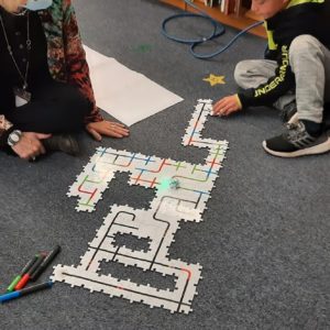 A Teacher And Student Sit On The Floor And Have Created A Path For A Robot To Follow That Is On Puzzles