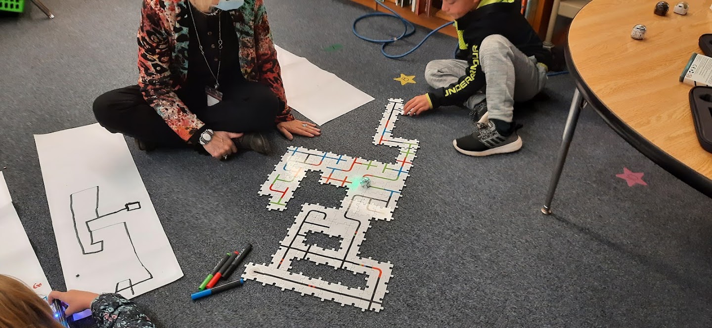a teacher and student sit on the floor and have created a path for a robot to follow that is on puzzles