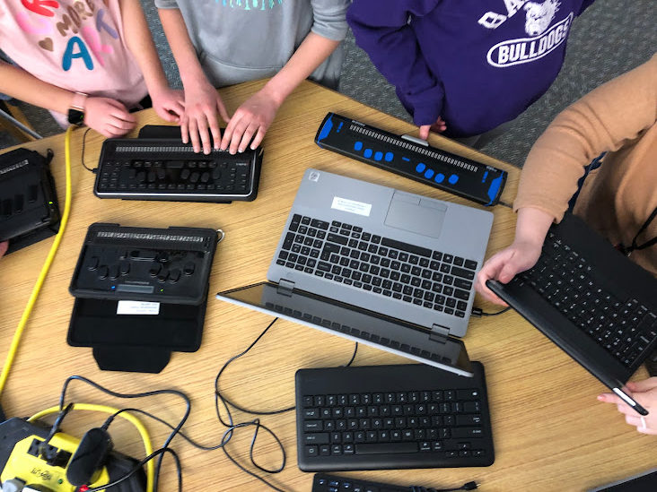hands touching braillenotes braille displays and keyboards spread out on a table