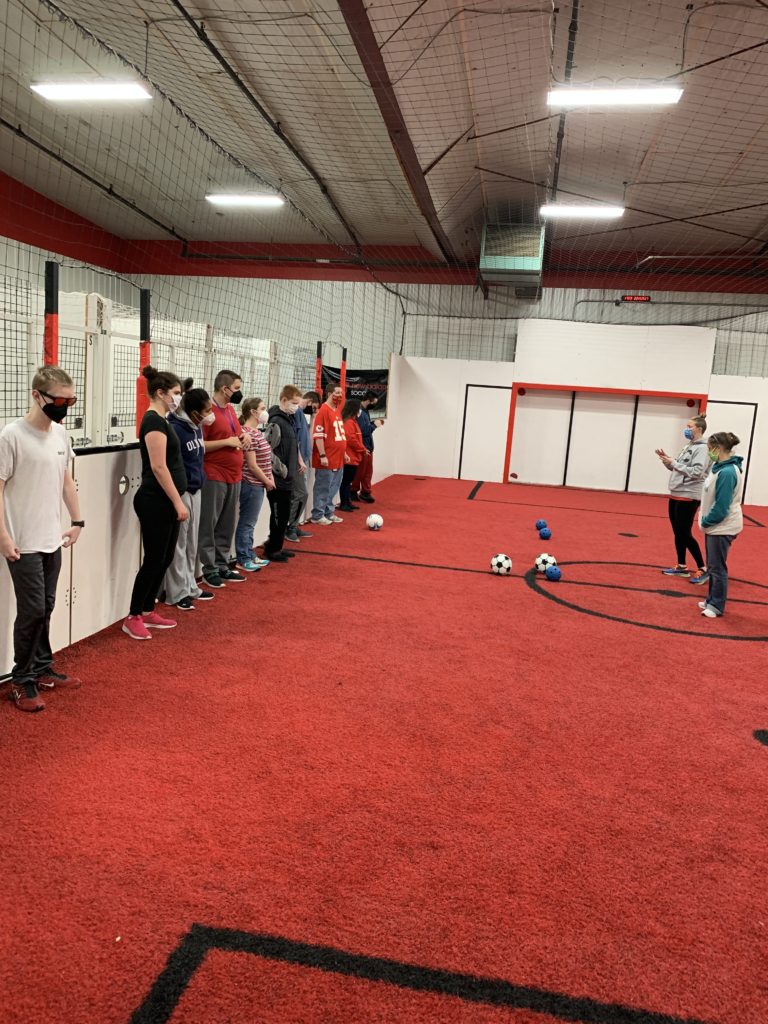 students stand against a wall on a soccer court with a red turf while 2 teachers stand in front of them giving instructions and a few soccer balls are on the turf