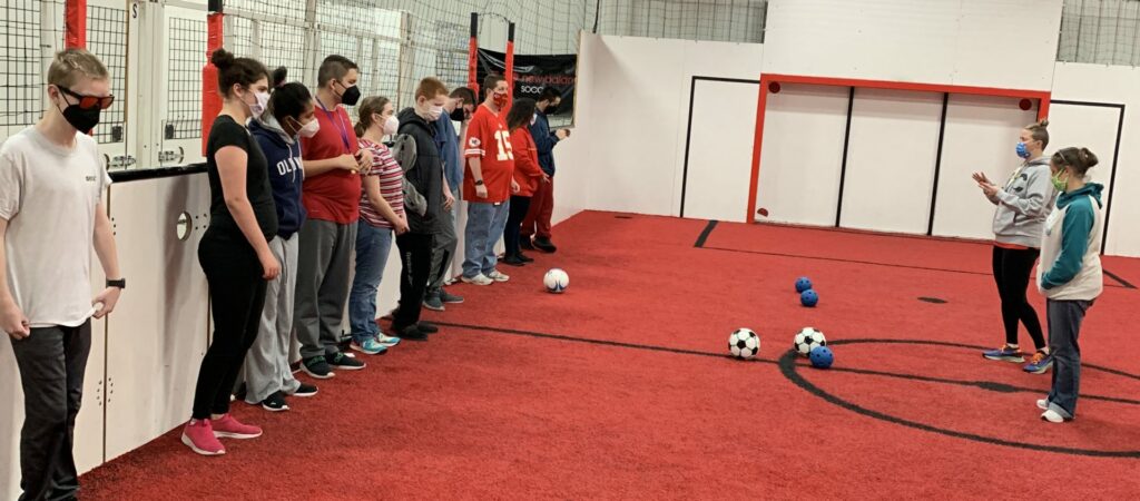 students stand against a wall on a soccer court with a red turf while 2 teachers stand in front of them giving instructions and a few soccer balls are on the turf