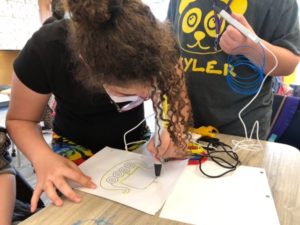 a girl with curly brown hair pulled into a bun leans over a table holding a 3D pen and drawing on a piece of paper