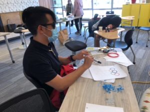 male student with short black hair and wearing sunglasses sits at a table holding a 3D pen and a drawing of the olympic rings on a paper in front of him