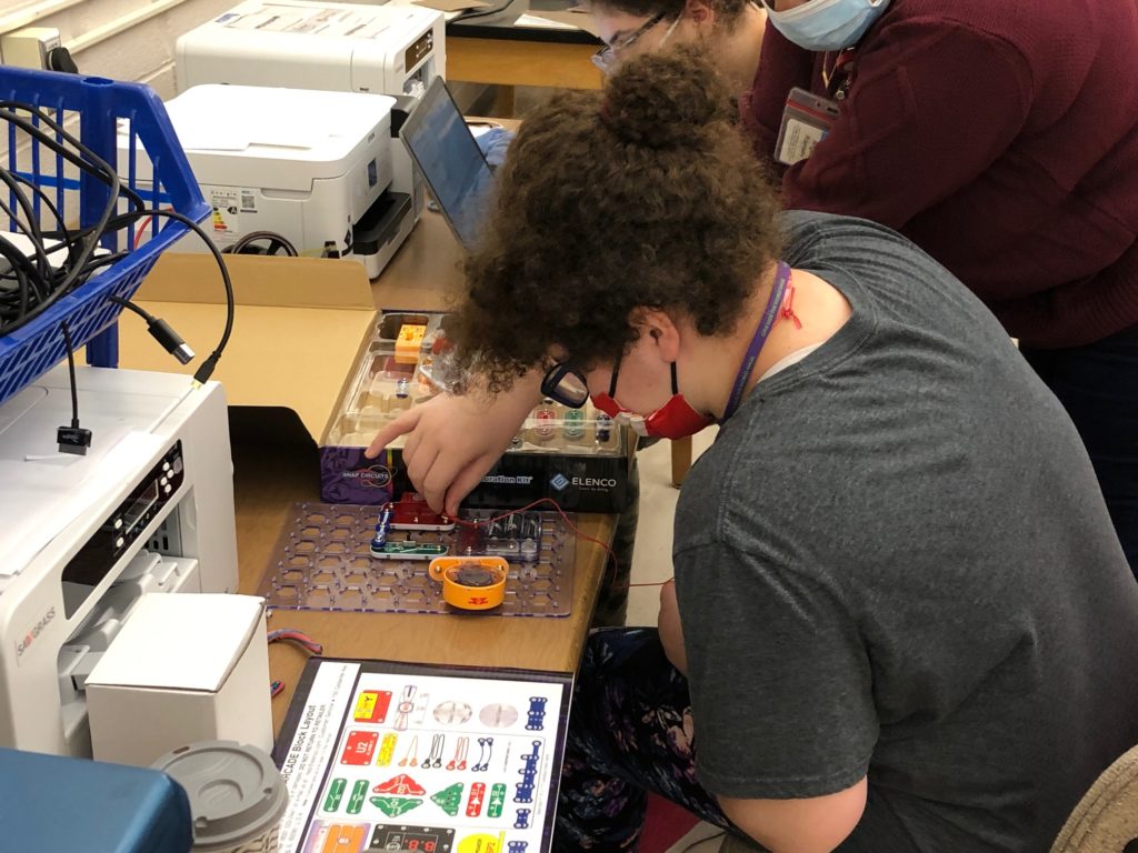 student with brown curly hair pulled into a bun and wearing glasses sits at a table working with snap circuits