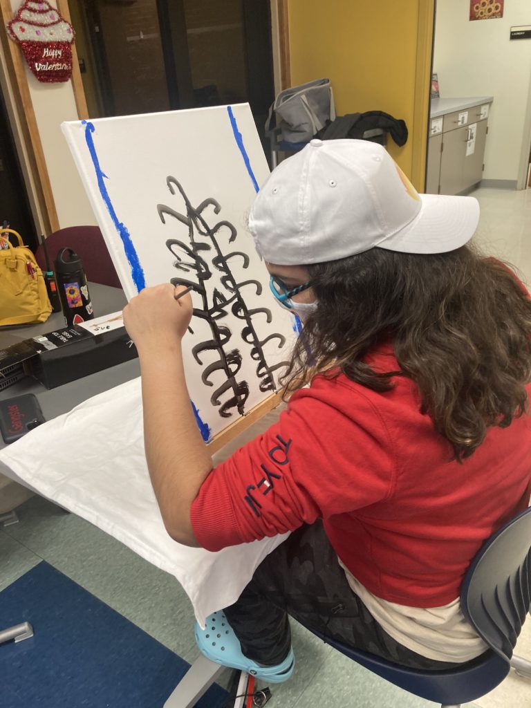 student with long brown hair wearing glasses and a baseball cap is painting on a canvas on an easel in front of her