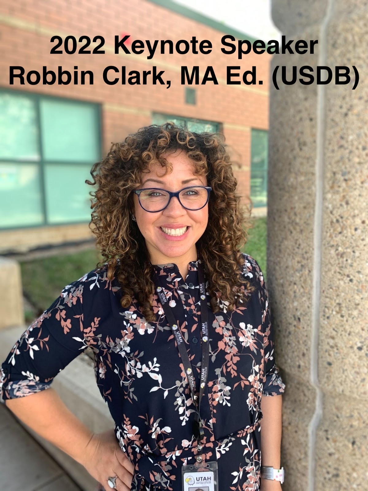 2022 Keynote Speaker Robbin Clark (MA Ed. USDB) stands against a pole wearing a dress with flowers, glasses, and has brown curly hair and smiles for the camera