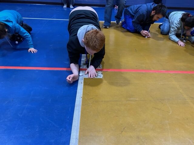 a red headed boy, knees on the floor, using a straw to blow a cotton ball across the gym floor
