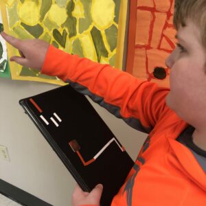 A student wearing a bright orange jacket holds a tactile map and points forward.