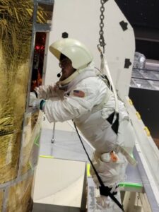 Tyler is performing the duties of a Mission Specialist and repairs a satellite while wearing a white space suit