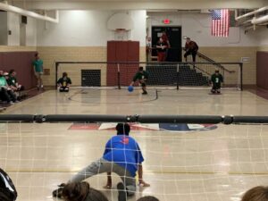 Students playing goalball in a gym, one student is crouched on the gym floor with a net behind him and on the opposite side of the gym is another student crouched down and holding a ball with a net behind them.