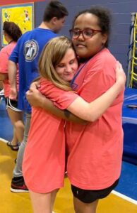 Two girls stand in the gym hugging and smiling.