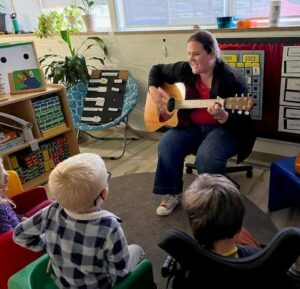 A female teacher plays guitar sitting down while small students sit on a carpet listening.