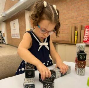 A young girl with pig tails and glasses plays with square robots on a table.