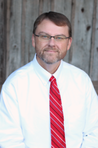 Dr. Derrick Smith - A Middle Aged Man With Short Brown Hair, Glasses And A Beard Smiles At The Camera Wearing A White Button Down Shirt And Red Tie.