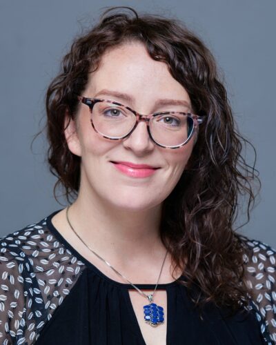 Heather Pedersen smiling for the camera with shoulder length curly hair and wearing glasses