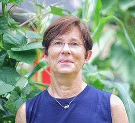 Judy Endicott - A Woman With Short Brown Hair And Rimless Glasses Smiles At The Camera Wearing A Blue Tank Top And Gold Necklace, Standing In Front Of Large Green Plants.
