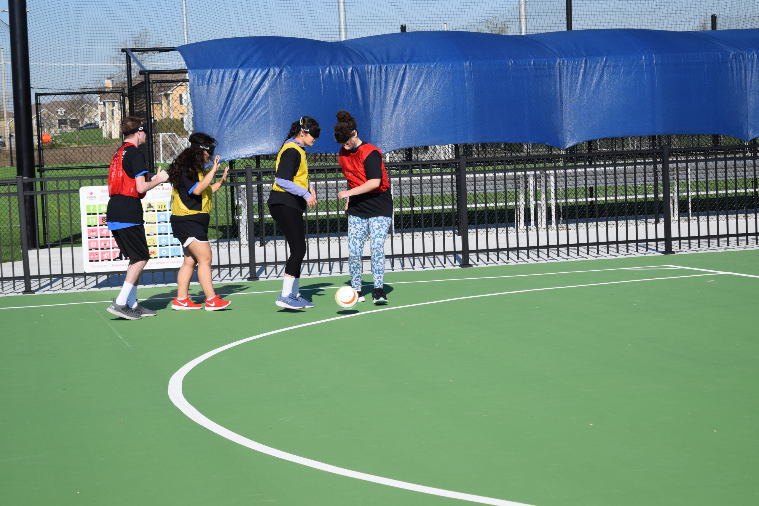 Four blindfolded participants, two in yellow shirts and two in red shirts, kick a soccer ball on a soccer field.