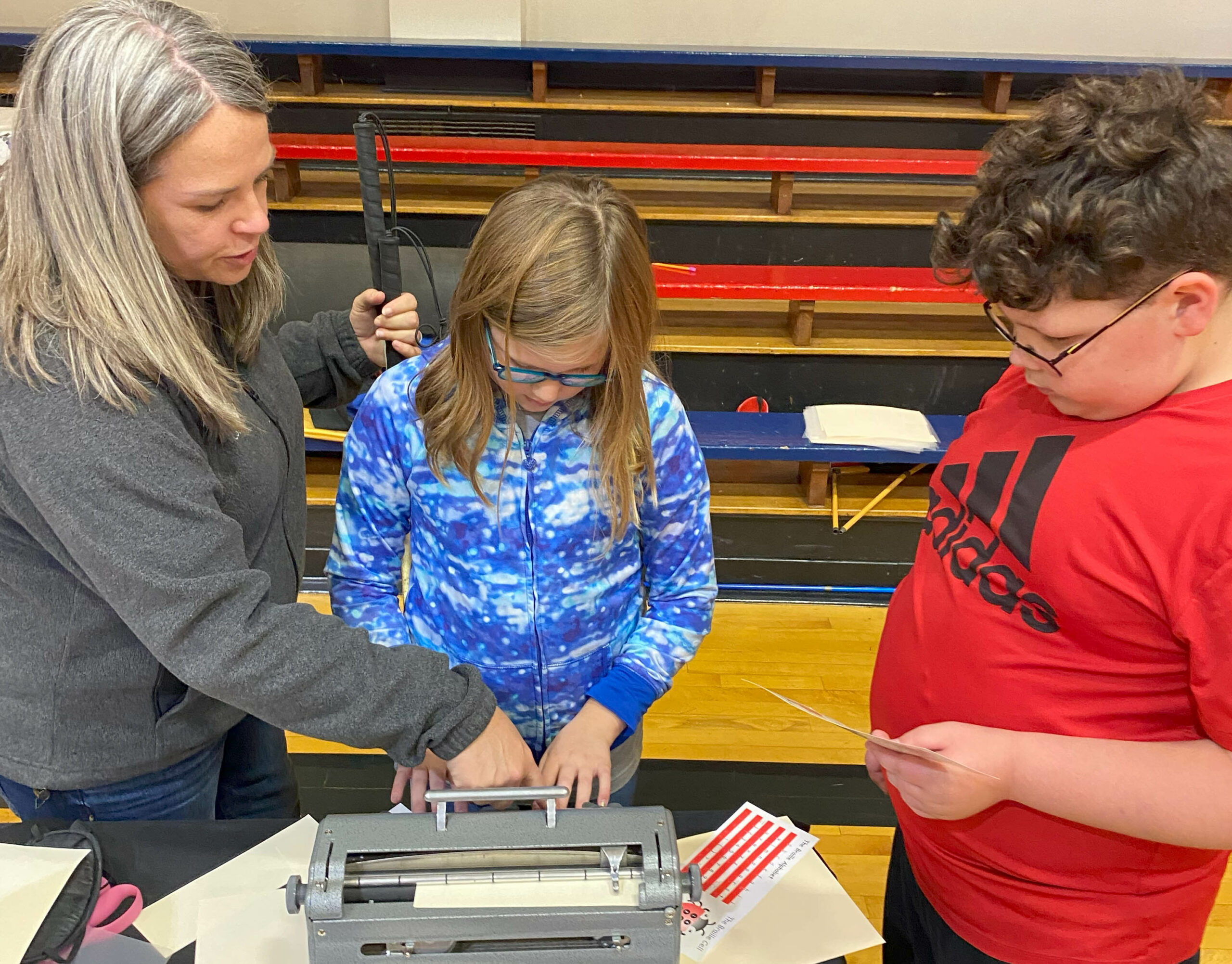 A woman with silver hair helps motion with her hand to a Perkins while a student uses both hands on the Perkins keys as another student looks on.