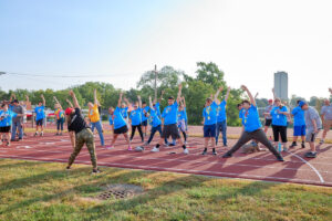 A group of students in blue shirts stand on the track with one arm extended overhead and stretch. They are being led by an adult who is standing in the grass.